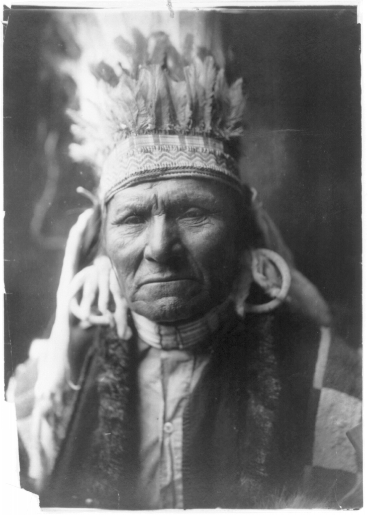 Native American chief wearing a feather headpiece, large earrings, and a vest