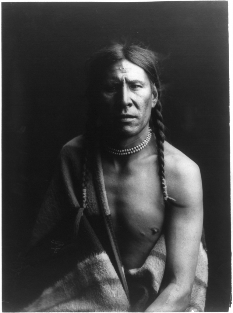 A Native American man with two long braids and a blanket covering half of his chest