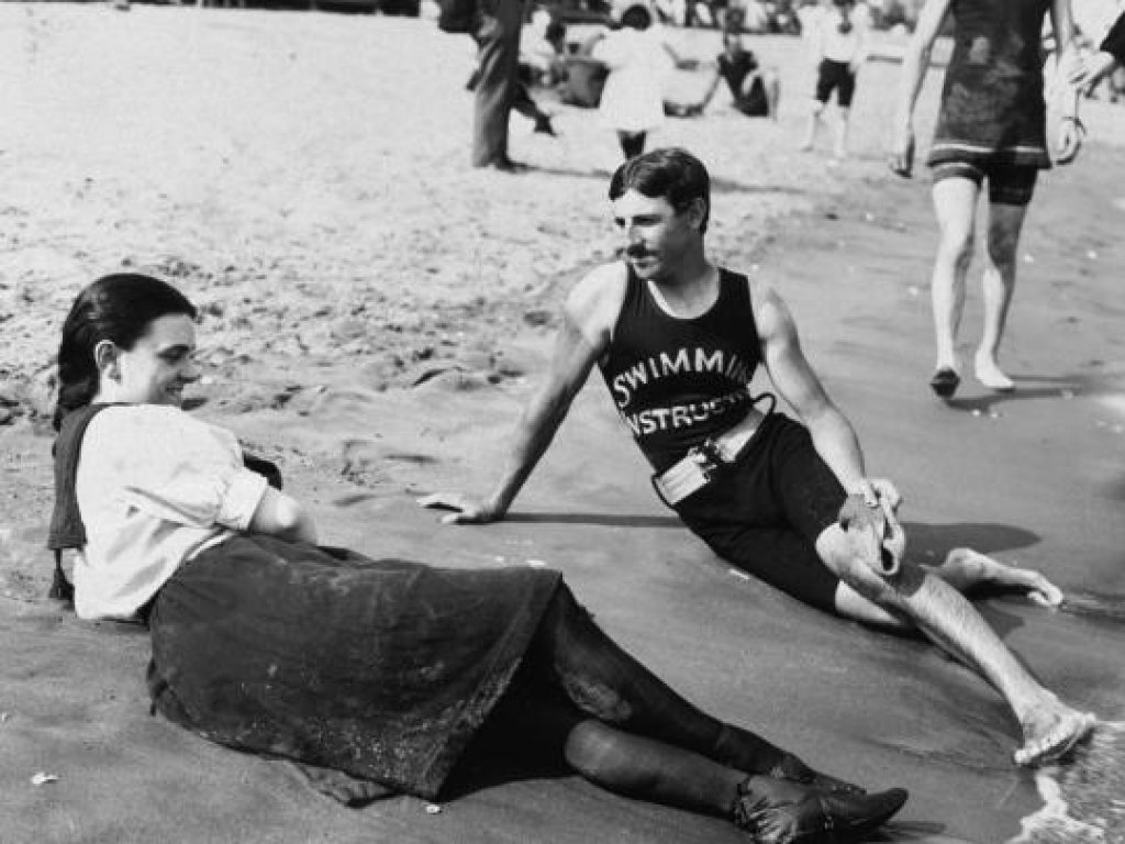 Young Nikola Tesla lays on the beach in a swimming costume talking to a young woman.