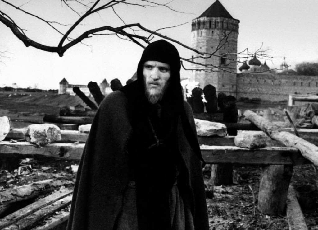 Man in cloak and hood stands in front of castle wall.
