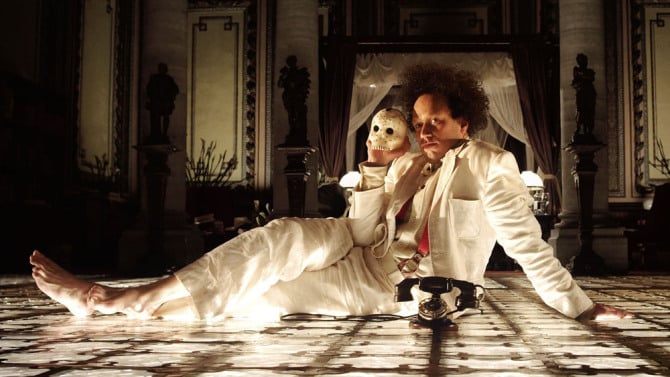 Peter Greenway dressed in a white suite, laying on the ground holding a skull