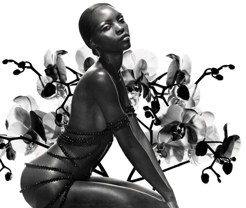 A black woman wrapped in jewels poses in front of a backdrop of orchids.
