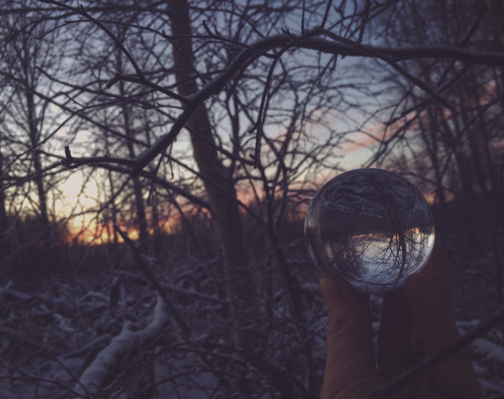 A hand holds a glass sphere in a winter forest at sunset.