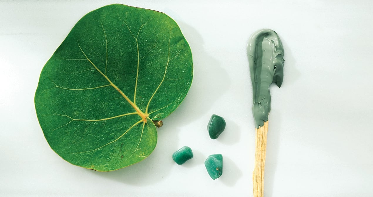 Large green sea-grape leaf lies on white with a wooden stick smeared with grey-green clay and three small green stones.