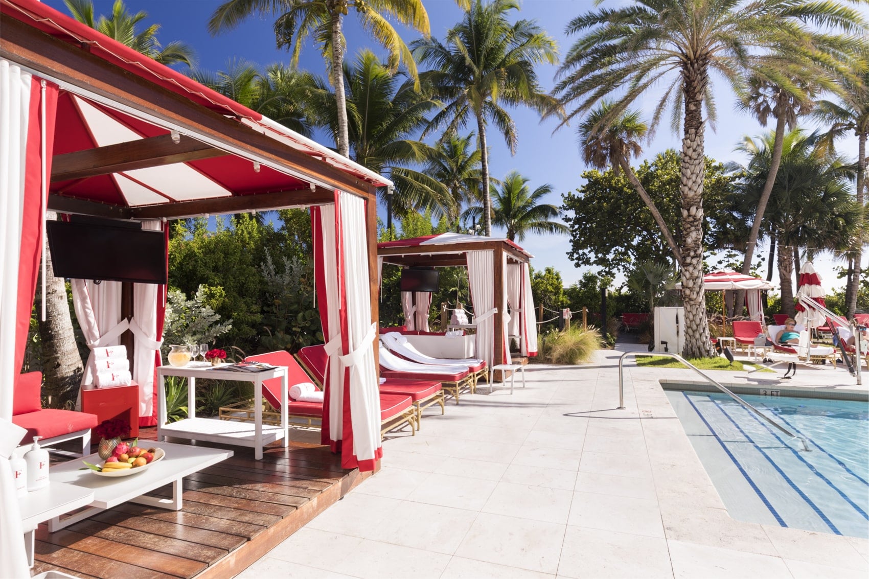 Under a red-and-white canopy, red cushions and a table with fruit and drinks sit inside a poolside cabana with a wall-mounted television.