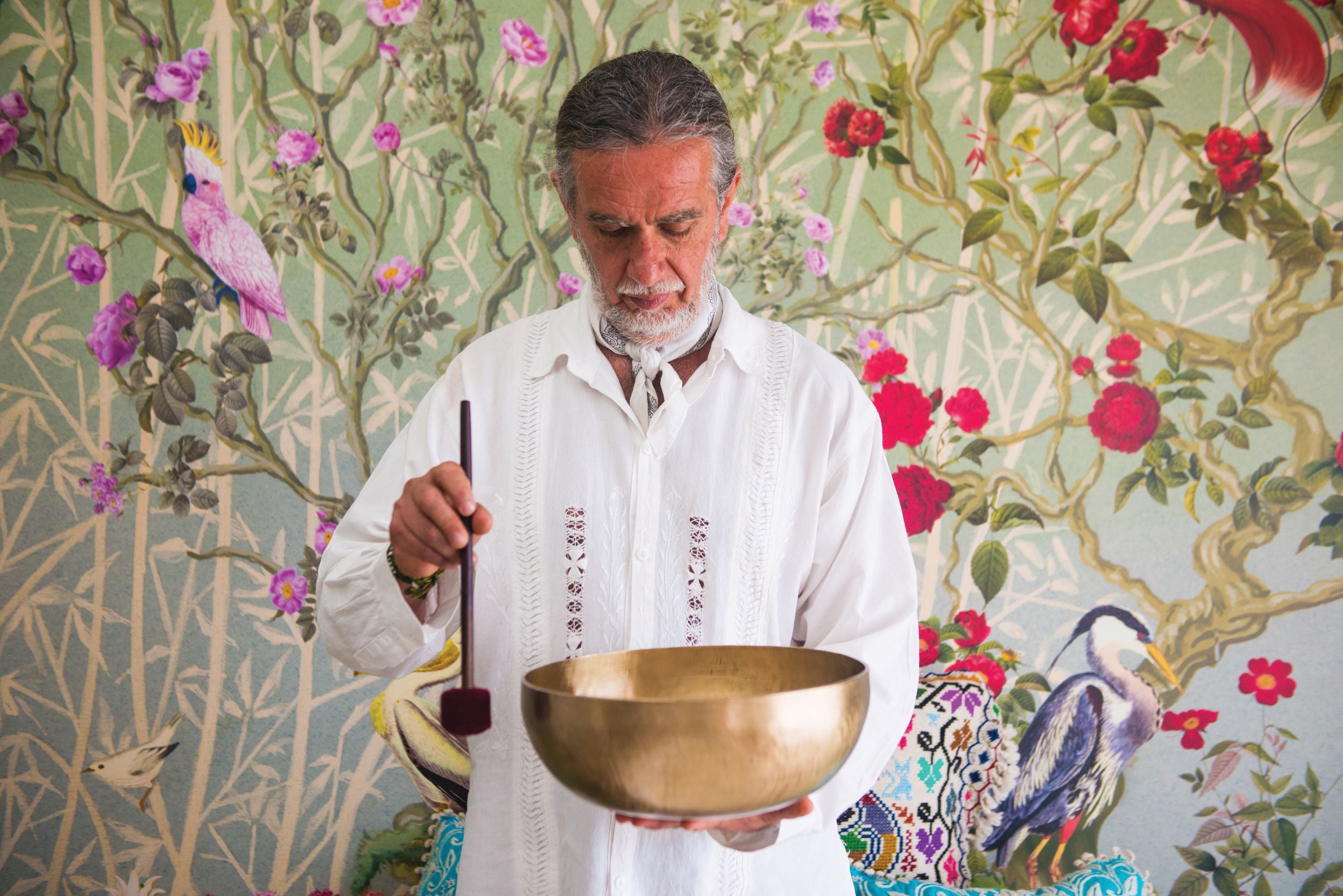 Green wallpaper with pink birds, red flowers and a blue heron frames a man in a white embroidered shirt looking down at a brass singing bowl.