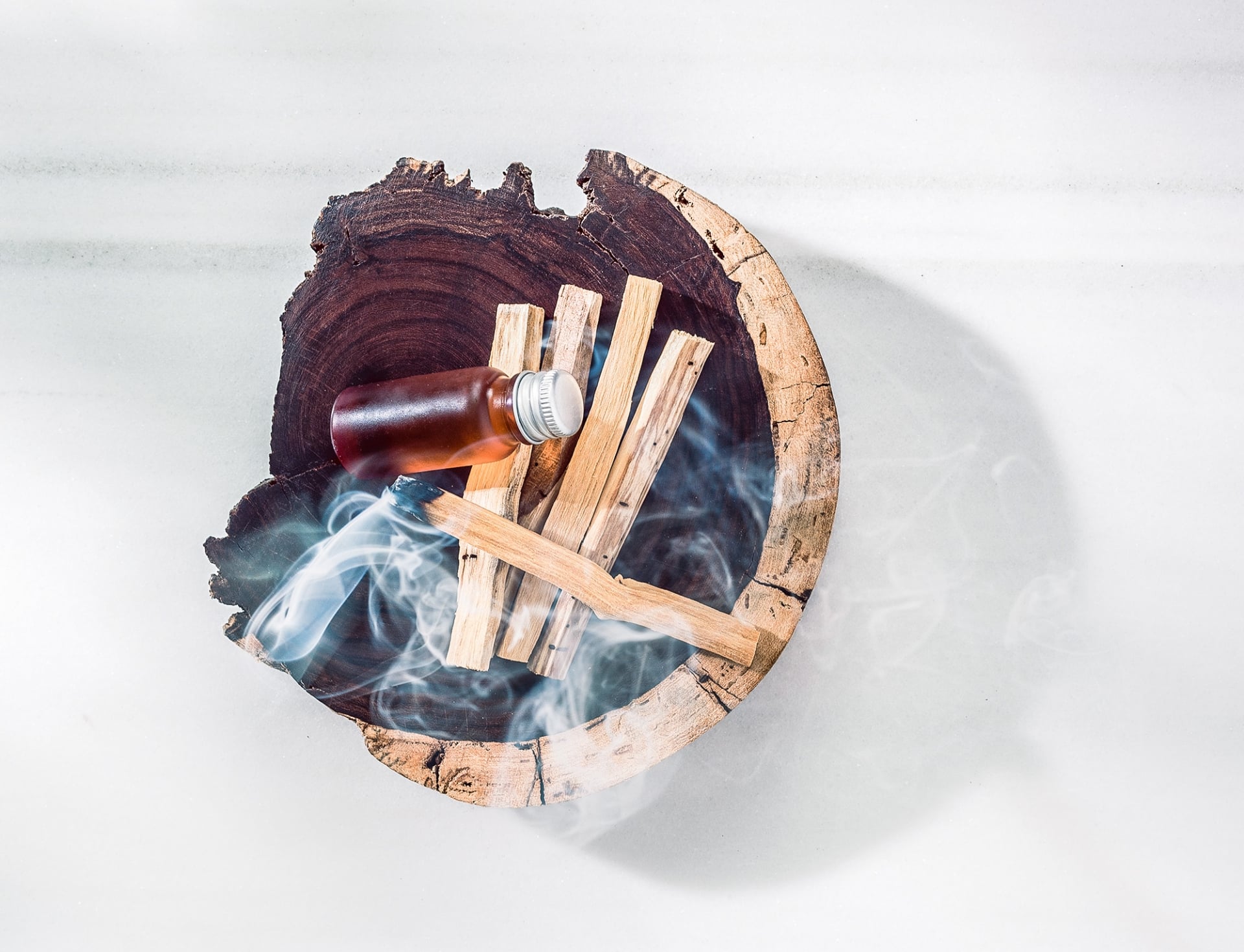 Palo santo sticks in a round wooden bowl, one smoking, sit beside a small amber glass bottle on white linen.