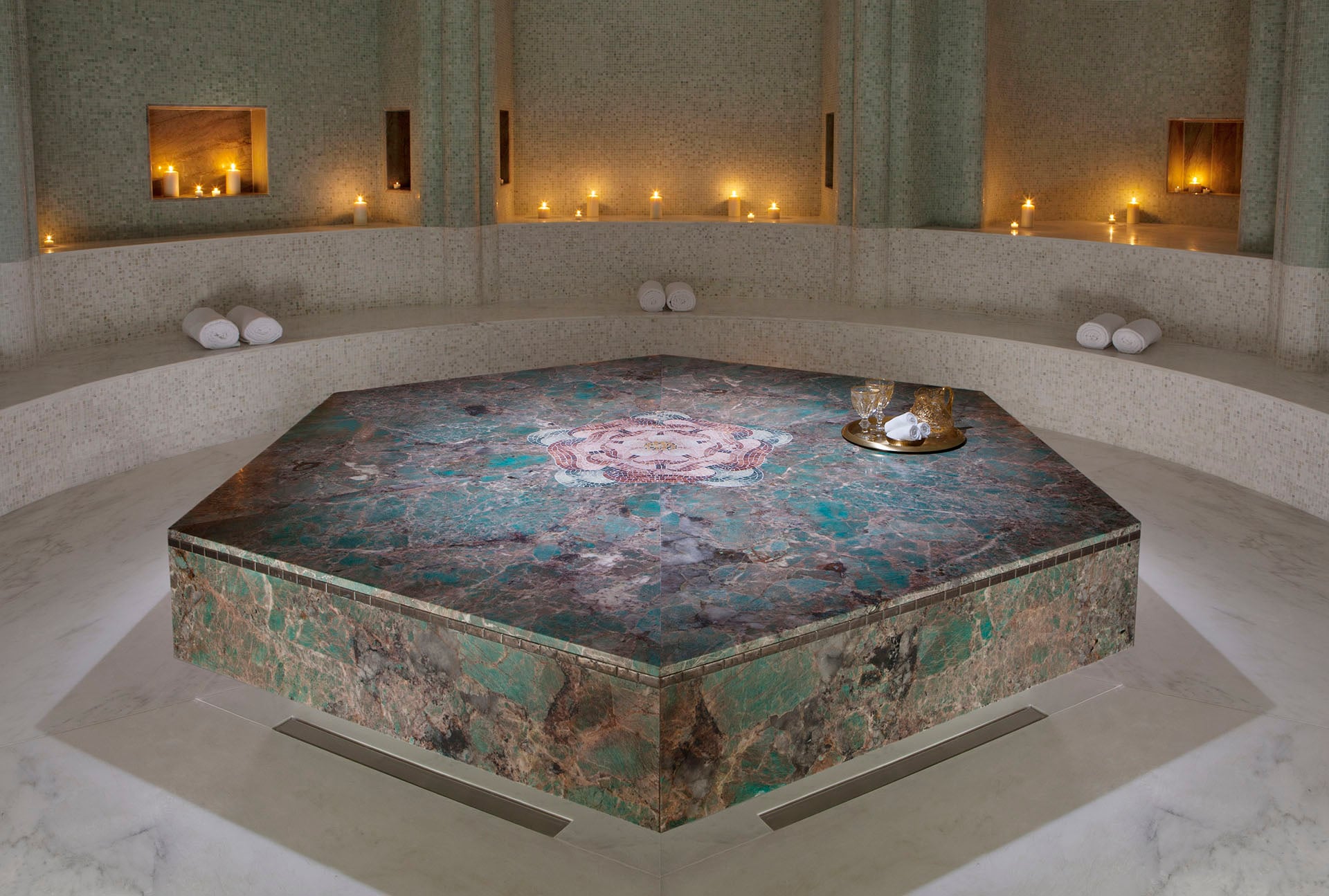 Candlelit room in Tierra Santa Healing House with a veined stone platform, a curved bench and candle niches in green mosaic walls.
