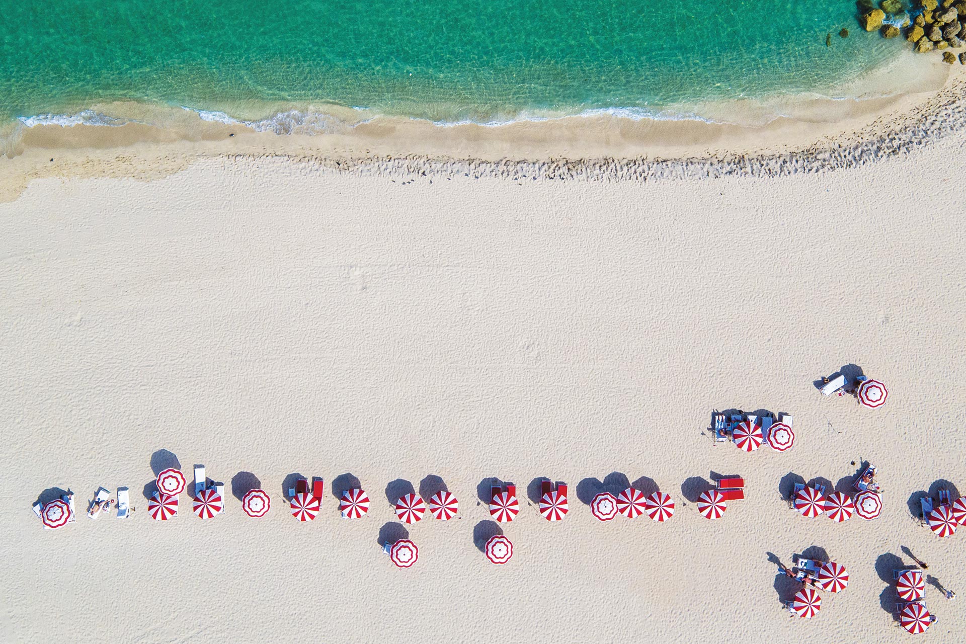 Umbrellas with red-and-white stripes line a white sand beach with turquoise water and a rocky jetty.