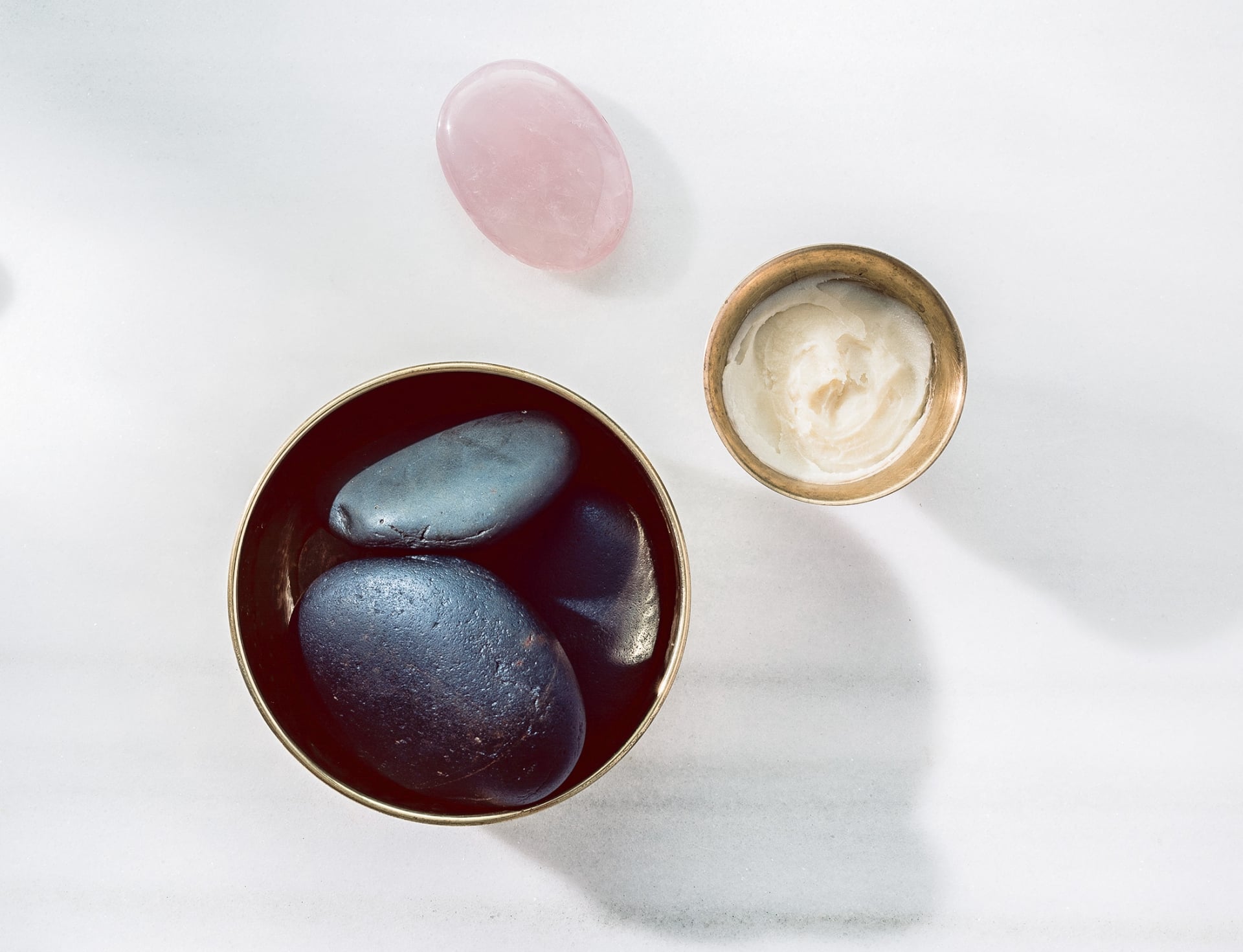 Smooth dark stones fill a brass bowl beside a pink rose-quartz stone and a small dish of cream on white marble.