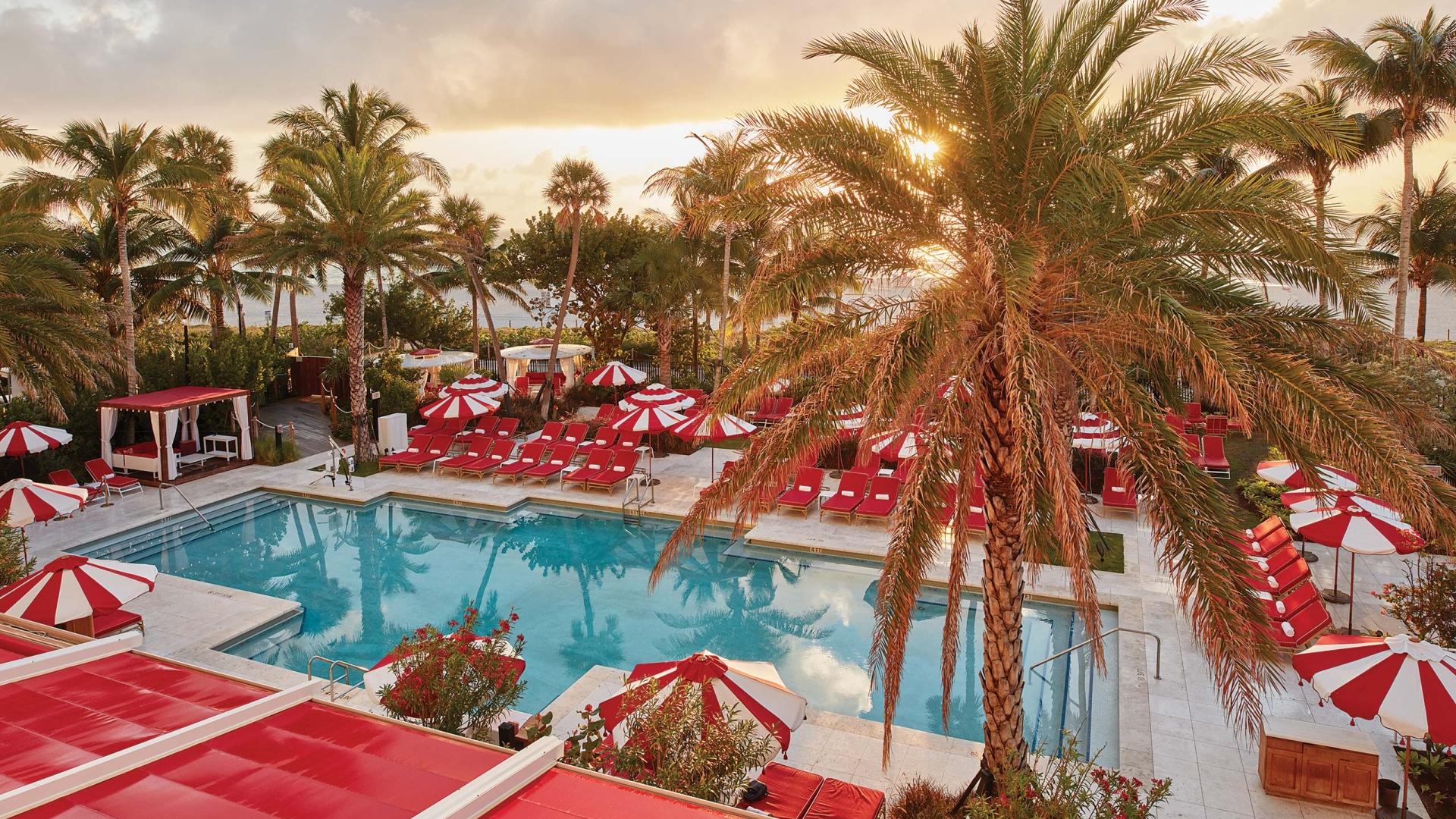 Aerial view of a pool area with red and white lounge chairs and umbrellas surrounded by palm trees at sunset.