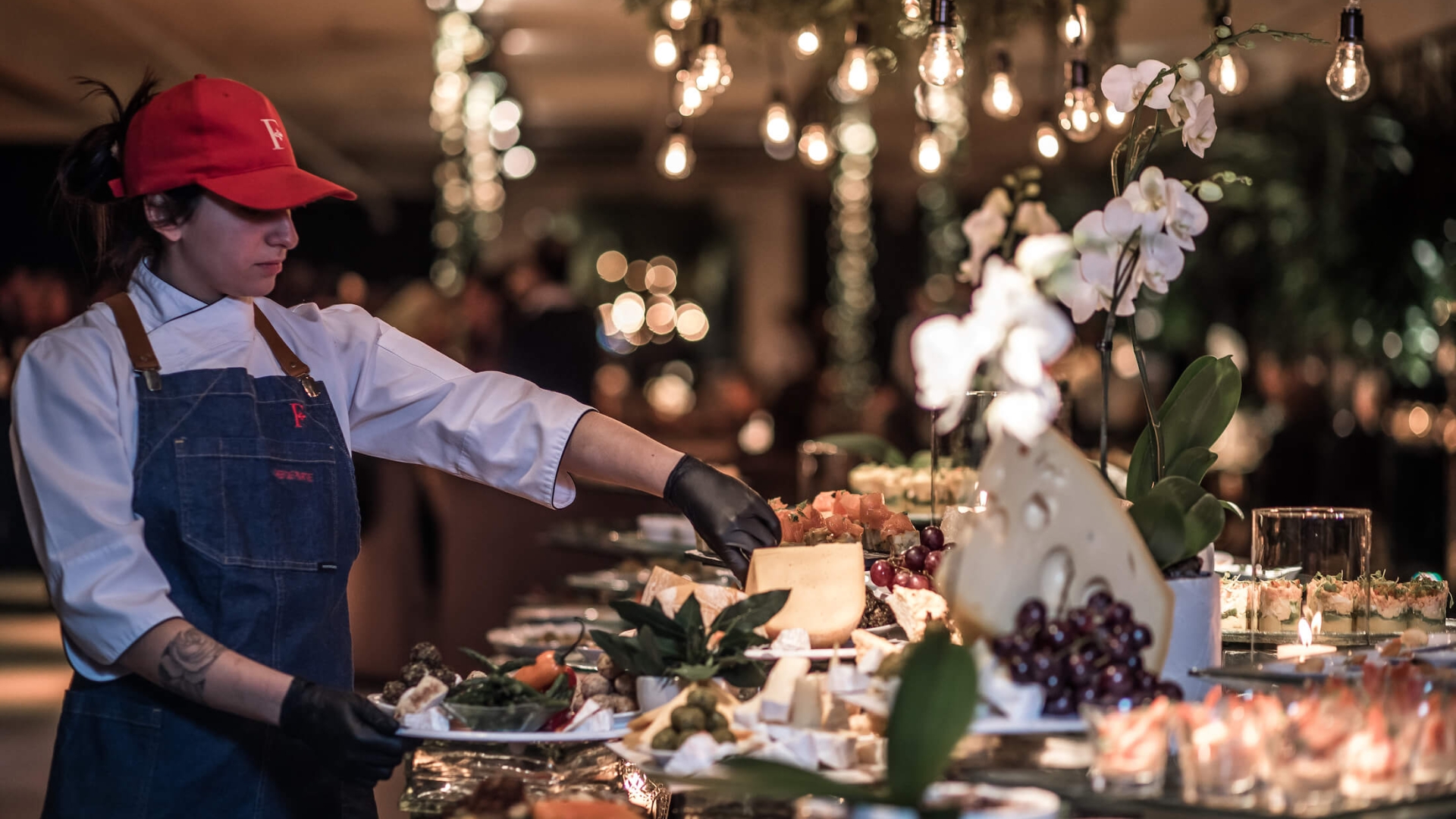 server wearing white shirt, blue apron, and red hat completes food prep on banquet table