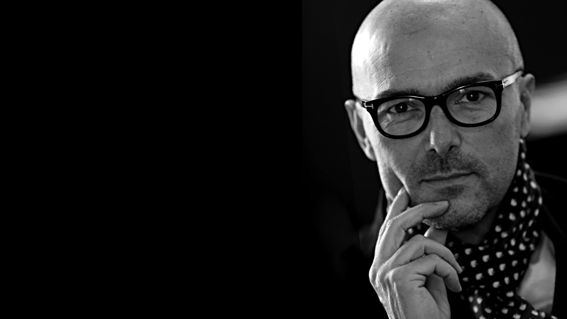 a bald man with glasses and polka dotted neck wrap stares at camera