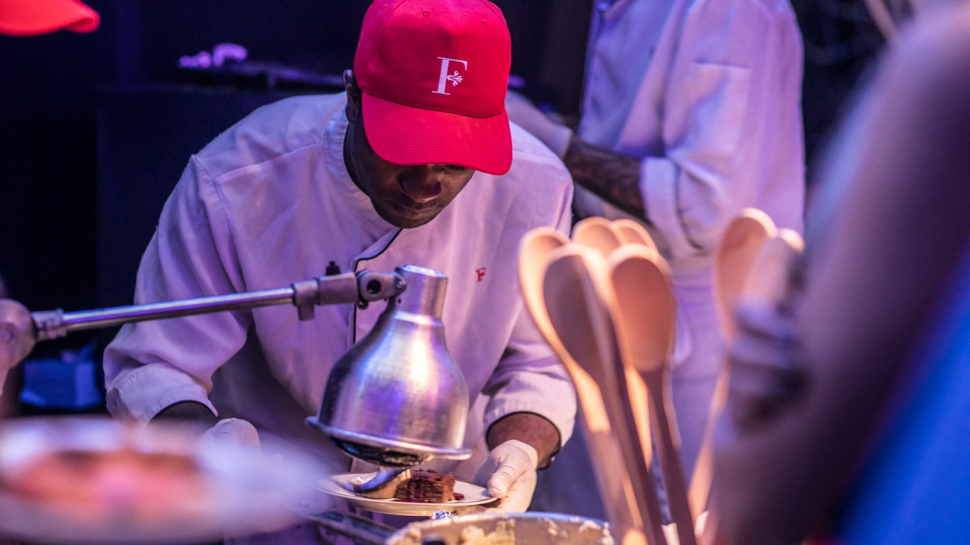 cook in white coat and red hat with embossed F looks down while preparing food
