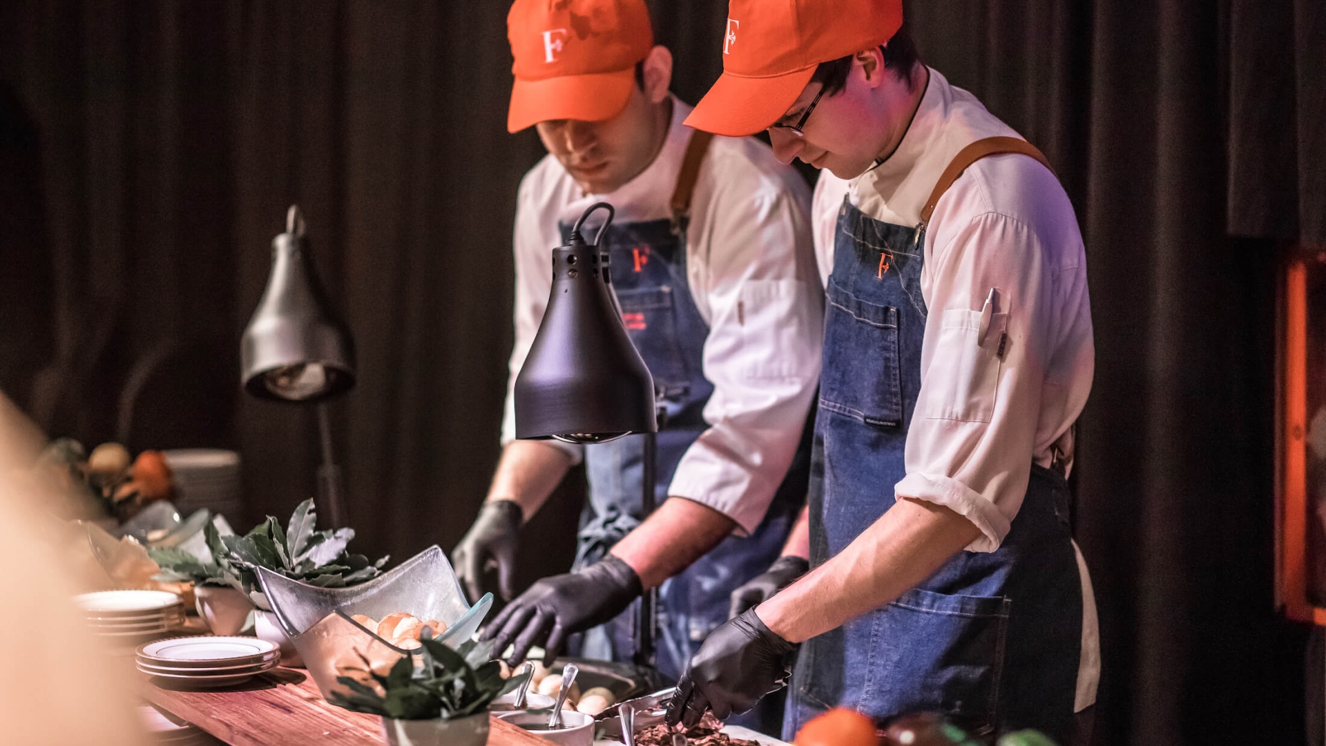 two servers wearing white shirts, blue aprons and red hats working on food preparation