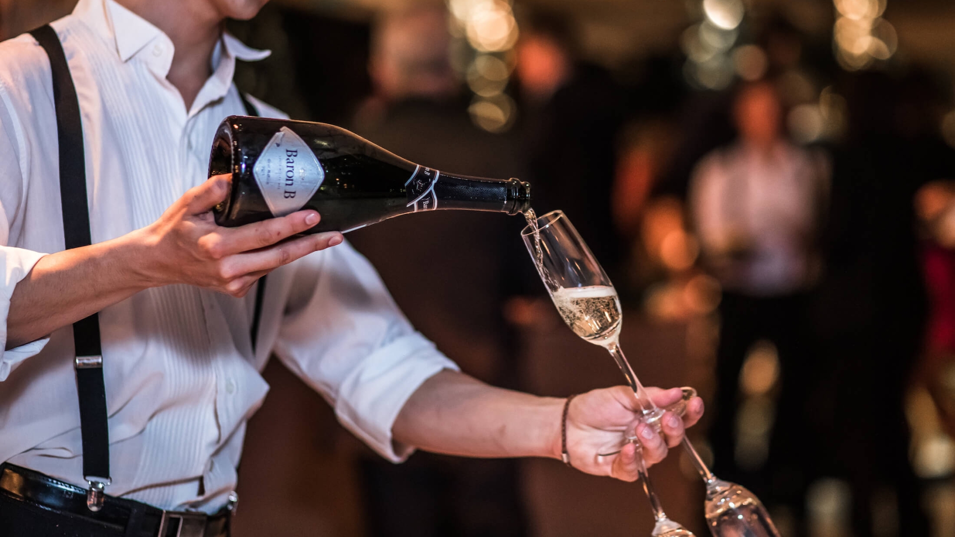server in white shirt and suspenders pours open bottle of sparkling wine into wine flute