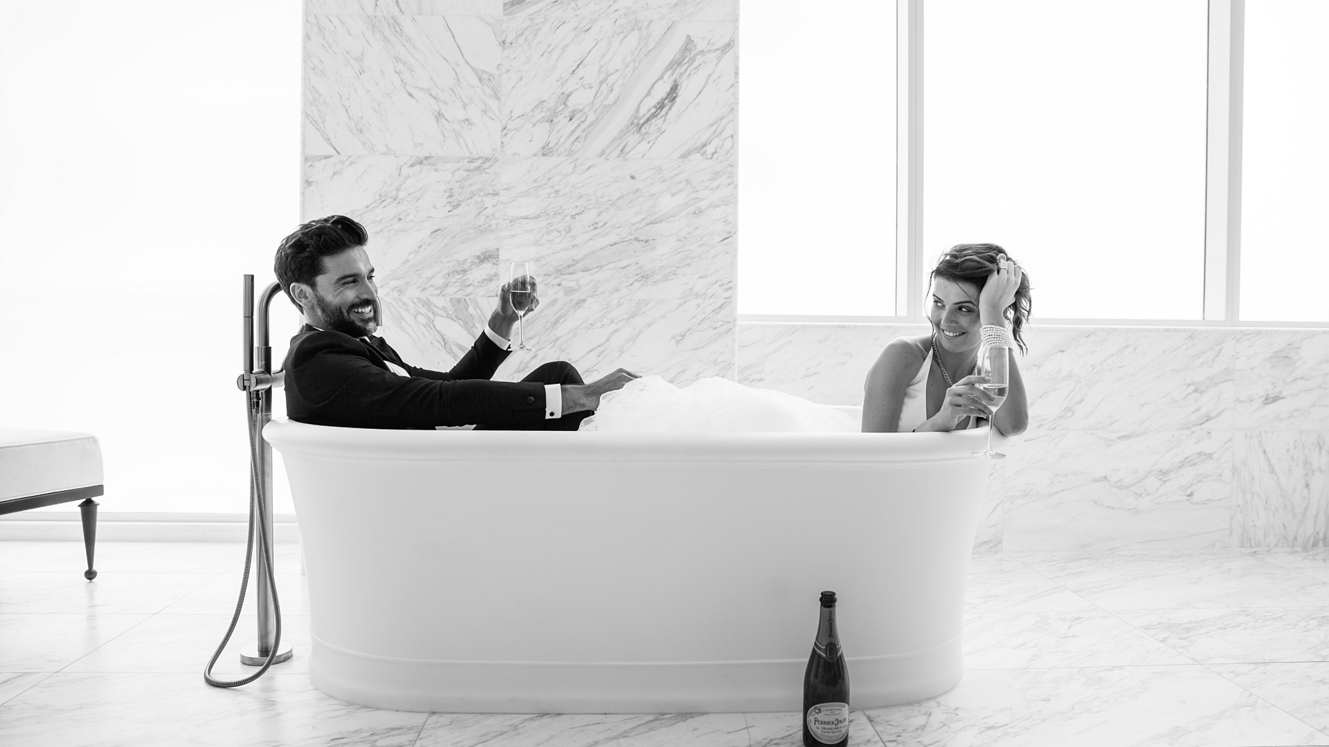 A black and white photo of a man and woman in a bubble bath with wine glasses
