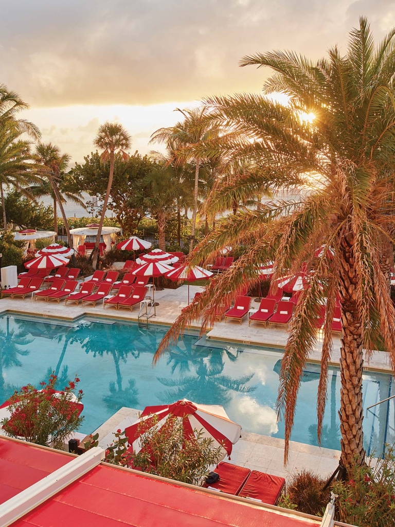 A poolside scene at sunset with palm trees, red lounge chairs, and red and white umbrellas.
