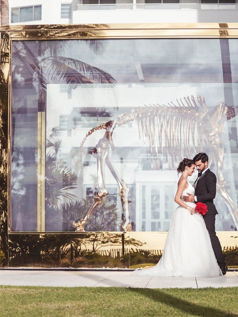 A large glass wall with a gold frame displays a dinosaur skeleton, with a couple in wedding attire standing in front.