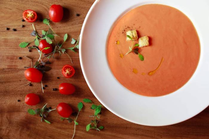 bowl of tomato soup sits on wooden table with sliced cherry tomatoes and greens lay beside it
