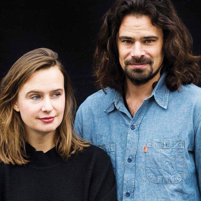 blonde haired woman and dark haired & bearded man stand side by side