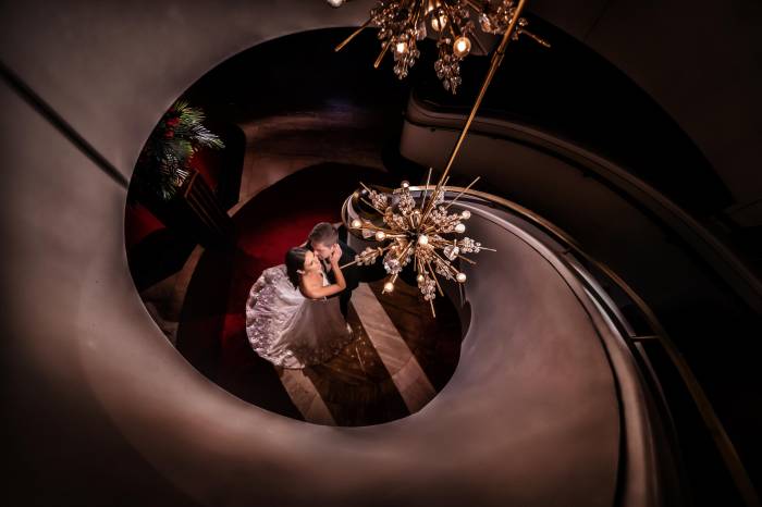 bride and groom stand at bottom of spiral staircase with chandelier hanging above them