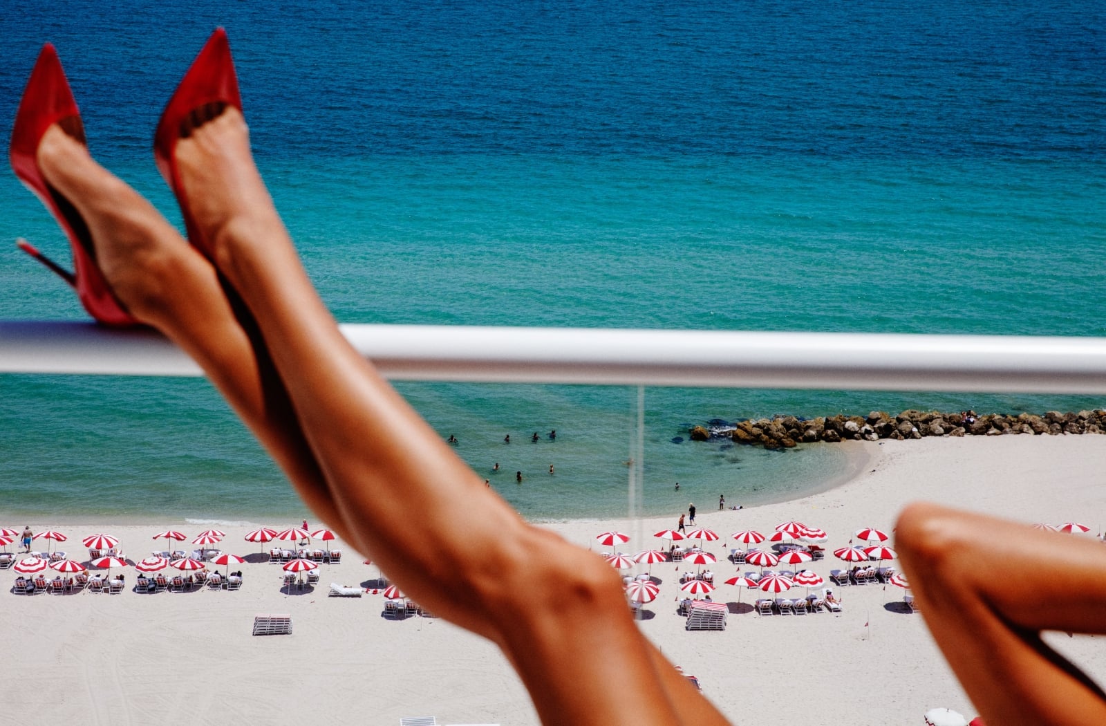 Woman in red heels overlooking Faena Miami Beach’s red-and-white umbrellas and turquoise ocean.