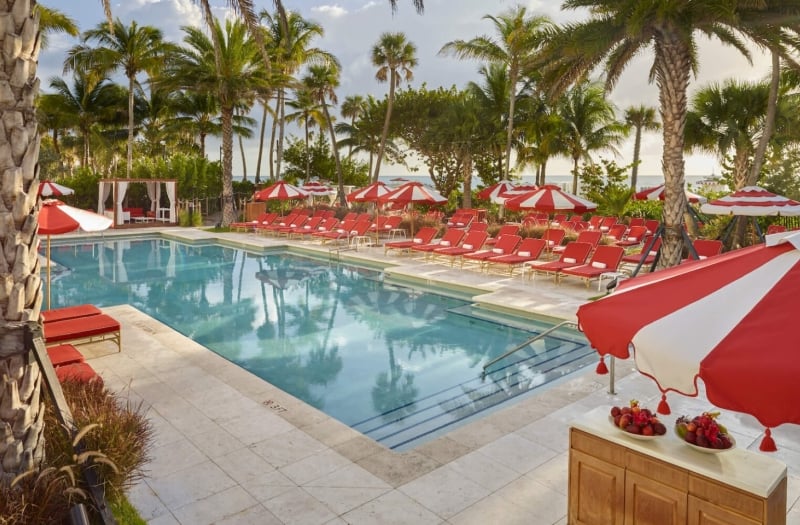 in-ground pool surrounded by red and white umbrellas and lounge chairs with palm trees in background