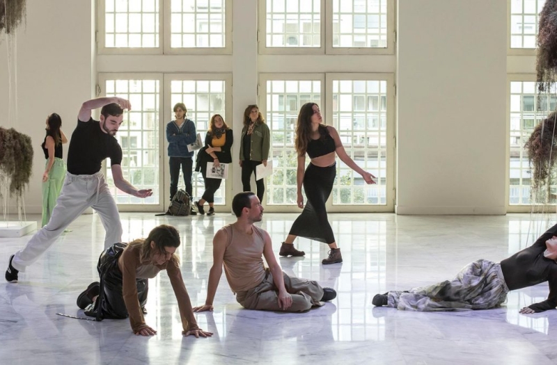 group of performers sit and stand in white walled room mid-performance