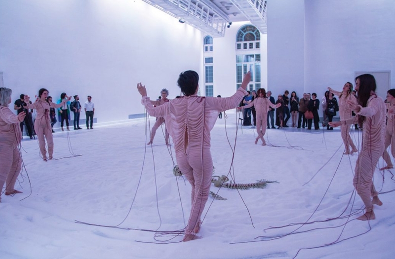 performers dressed in pale pink skin tight fabric dance on white cloth covering the floor