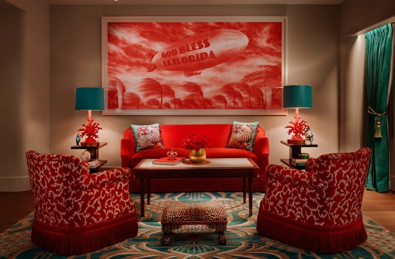 red chairs and couch sit on blue printed rug in front of painting of blimp that reads "God Bless La Florida"