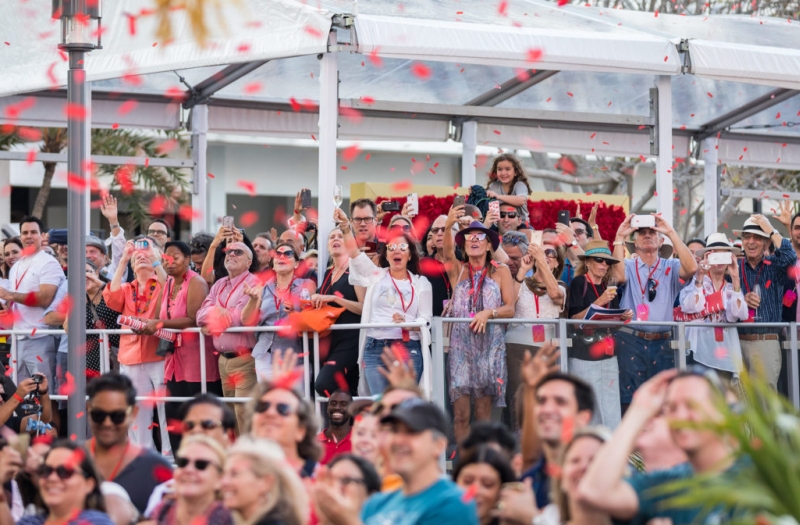 crowd stands along fence while cheering and waving with red confetti in the air