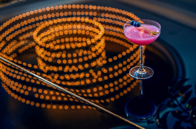 Pink cocktail with blueberry garnish on a piano with the reflection of a large chandelier 