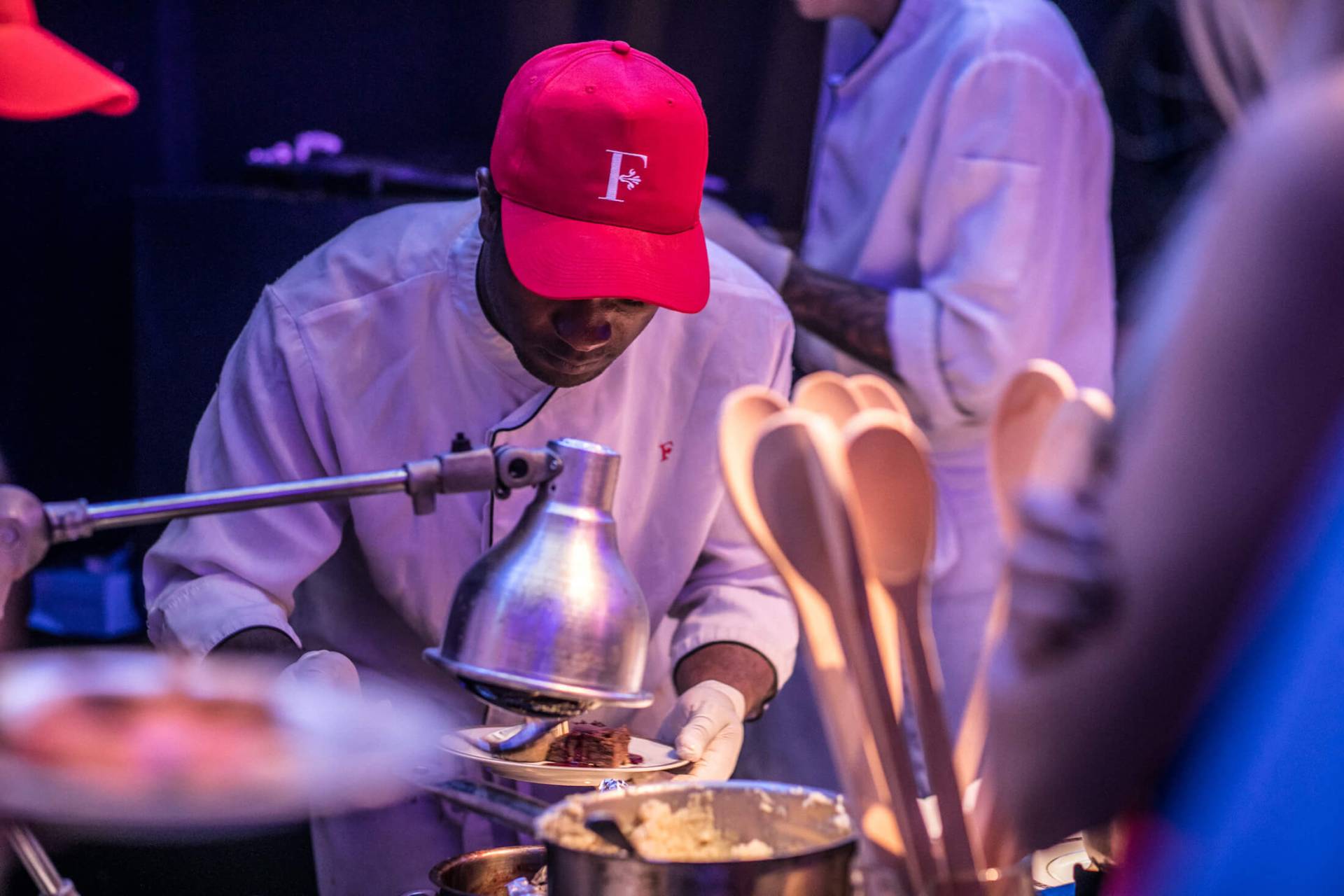 cook in white coat and red hat with embossed F looks down while preparing food