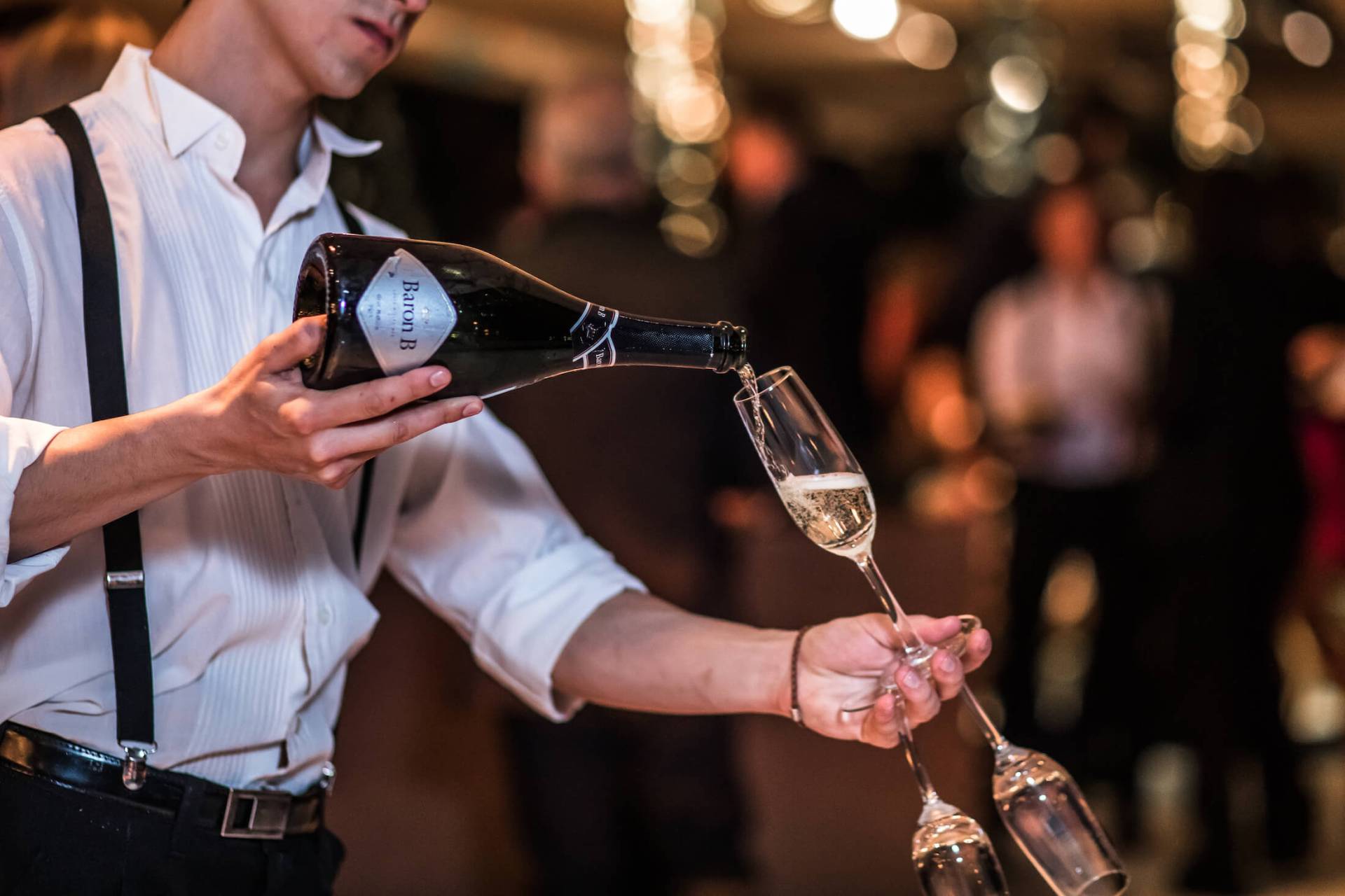 server in white shirt and suspenders pours open bottle of sparkling wine into wine flute