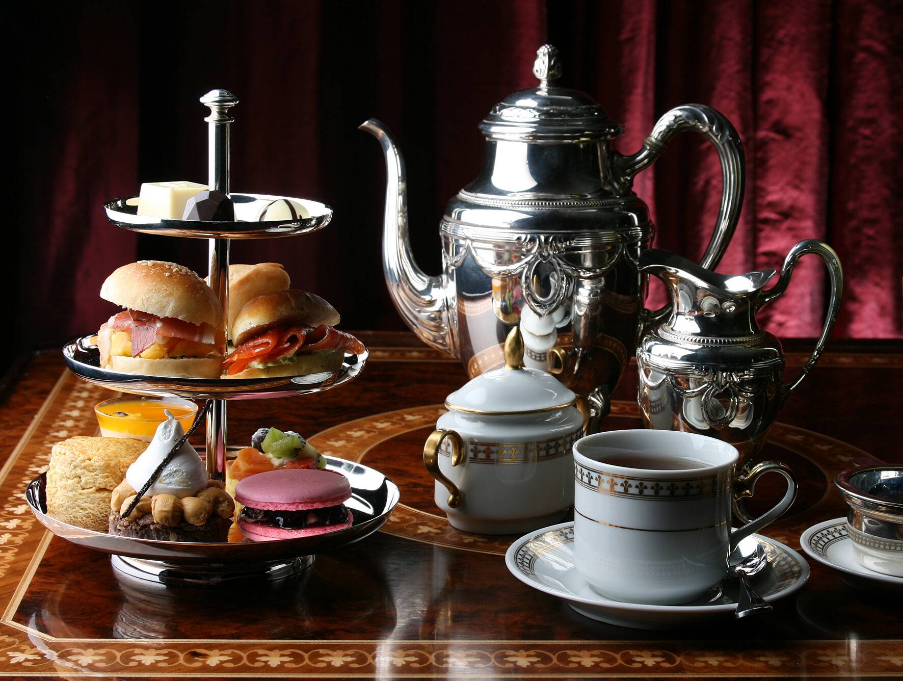silver teapot and milk jug sit on table beside two tea cups and a multi-tiered tray of sandwiches and desserts