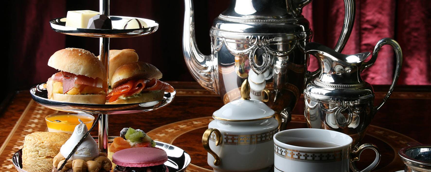 silver teapot and milk jug sit on table beside two tea cups and a multi-tiered tray of sandwiches and desserts