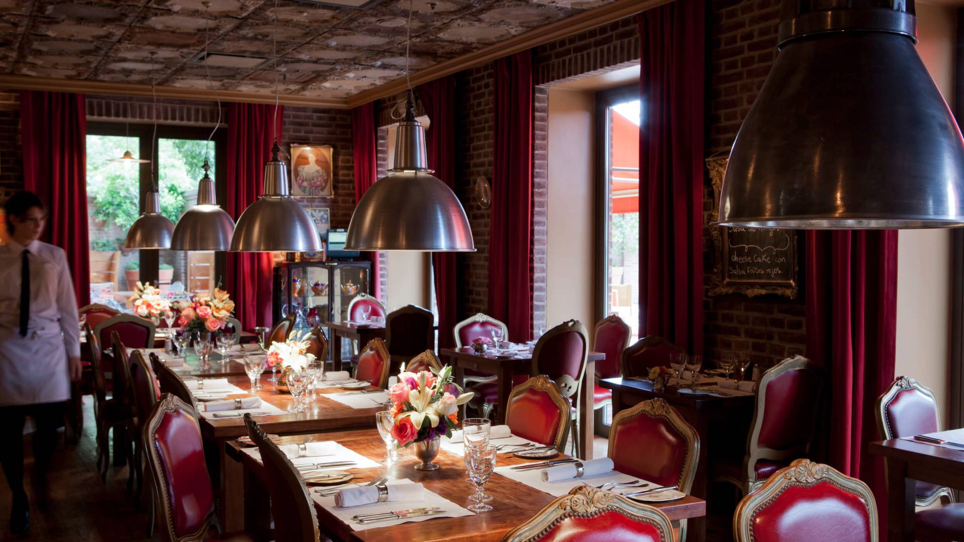 server walks alongside table in room with tin ceiling, large light fixtures, and red chairs framed in gold