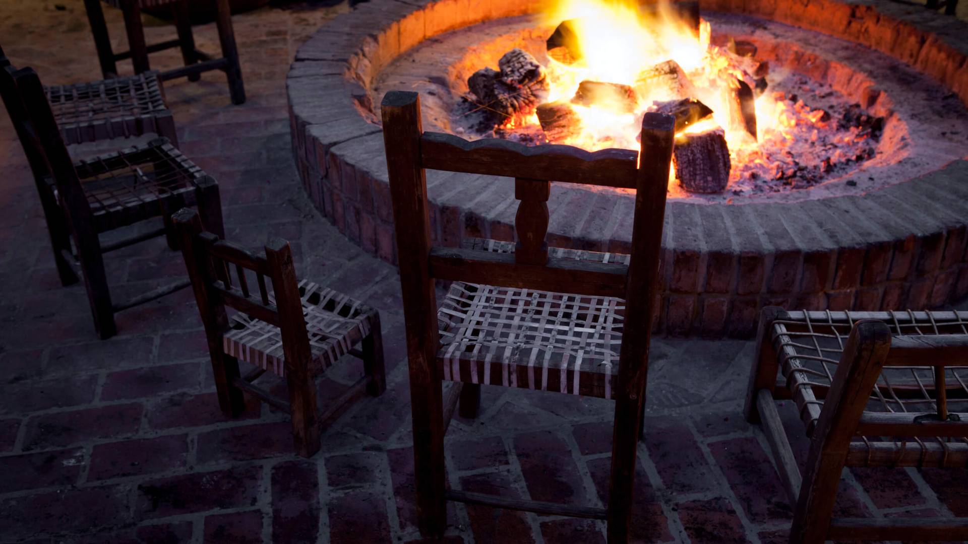 wooden chairs sit around a brick fireplace with small fire burning 