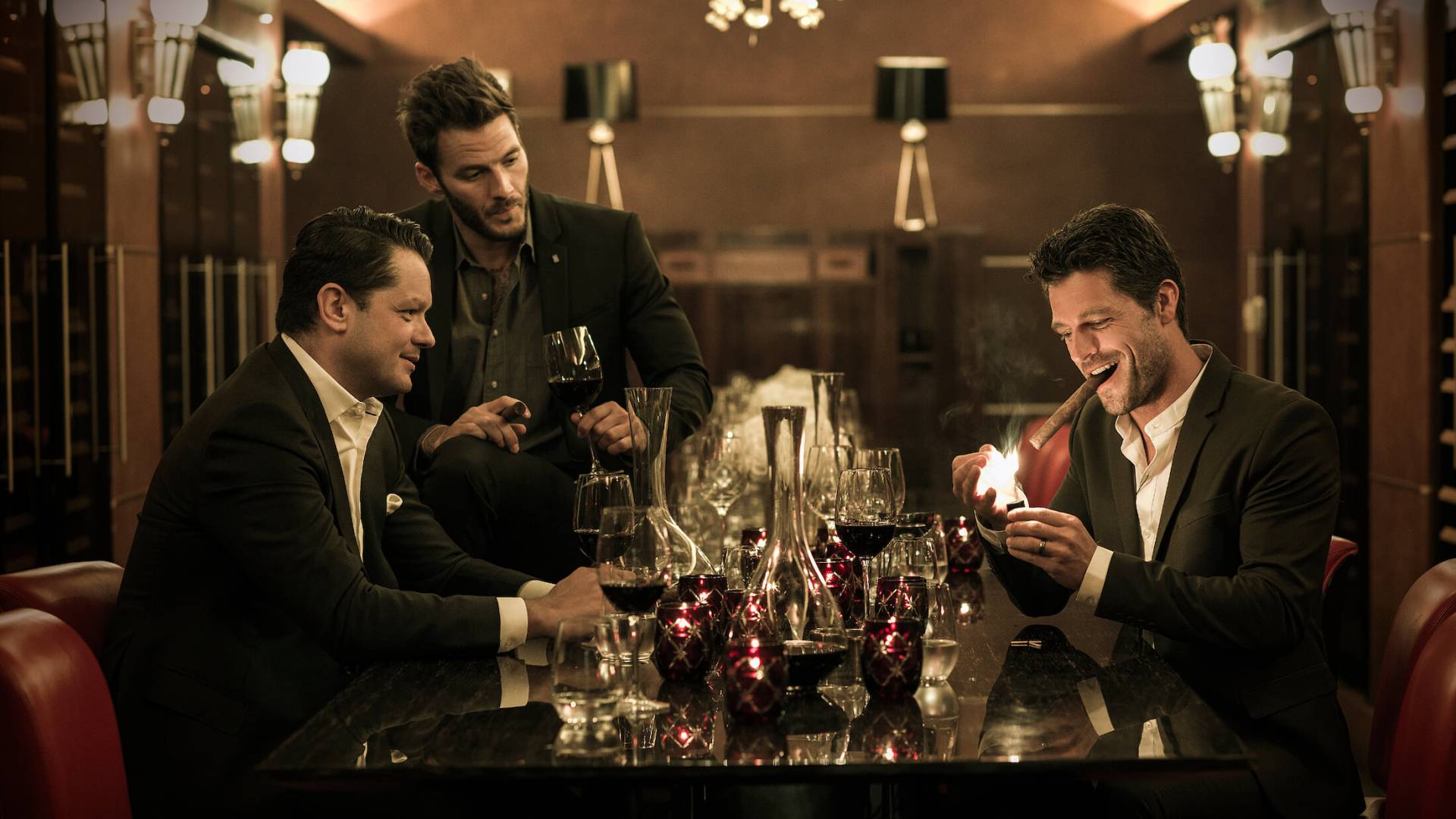 three men in black suits light up cigars sitting at table covered in glasses