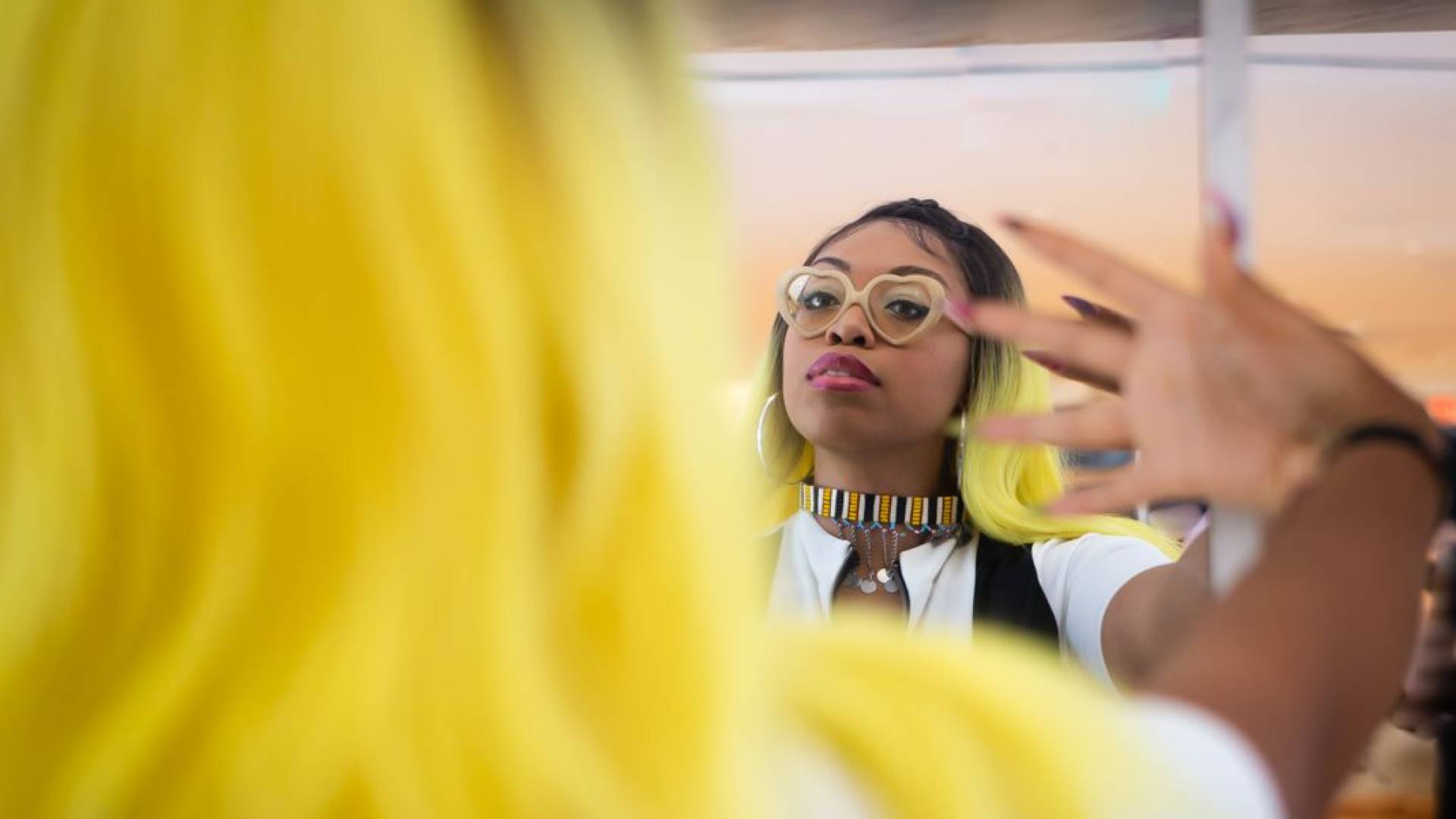 woman with yellow hair and glasses looks at herself in mirror with hand on glass