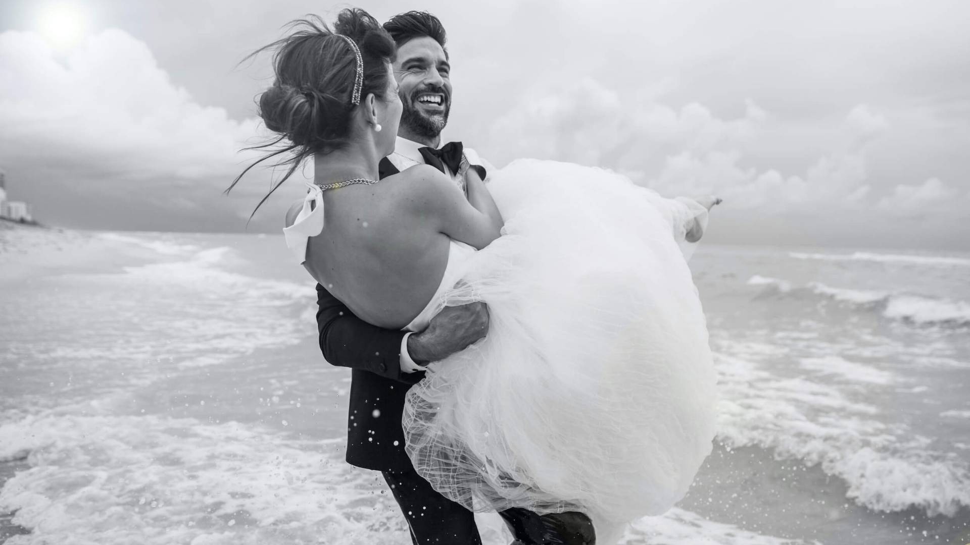 man in suit carries woman in white dress in his arms as he walks into ocean waves