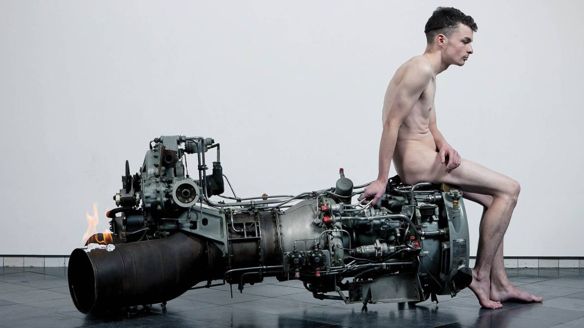 naked white man sits on the right edge of a large engine-like metal object