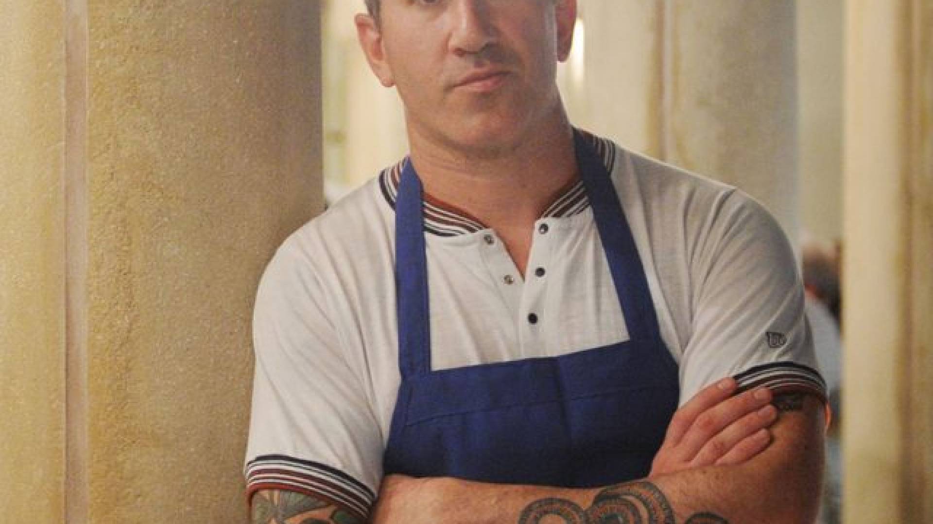 man with short hair leans on wall with arms crossed wearing chef's apron