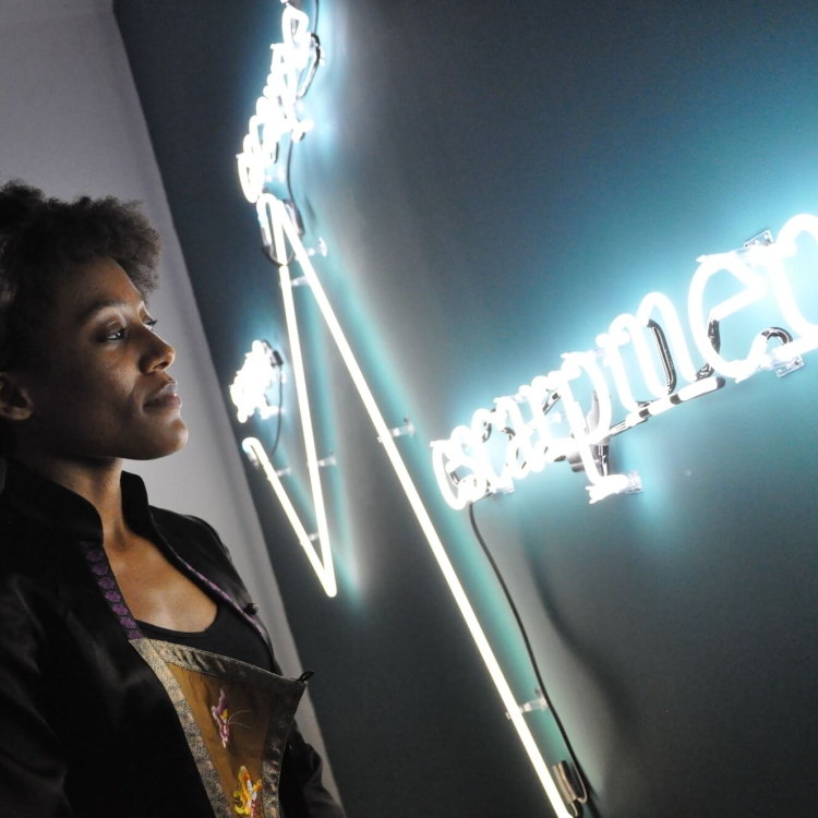 woman with hair pulled back looks closely at lit-up neon sign on wall