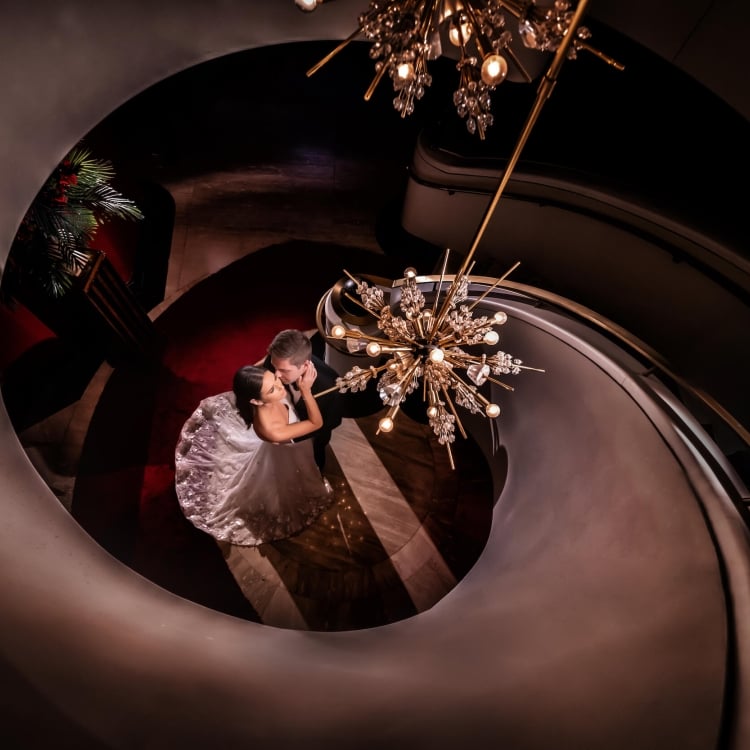 bride and groom stand at bottom of spiral staircase with chandelier hanging above them