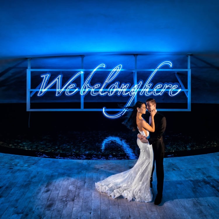 bride and groom stand in front of blue neon sign that reads "We belong here"