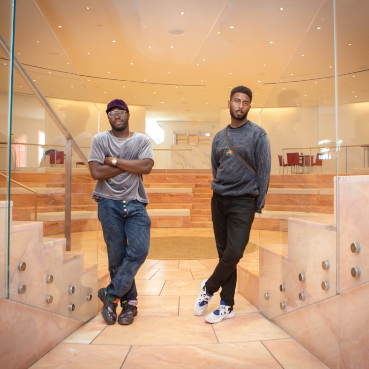 two men standing apart with one leg crossed in front of the other look toward camera
