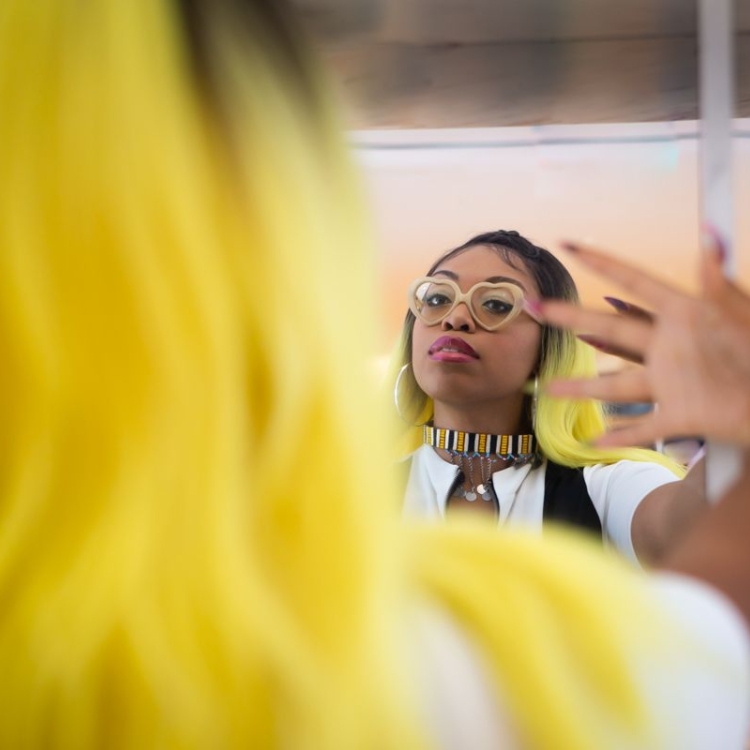 woman with yellow hair and glasses looks at herself in mirror with hand on glass