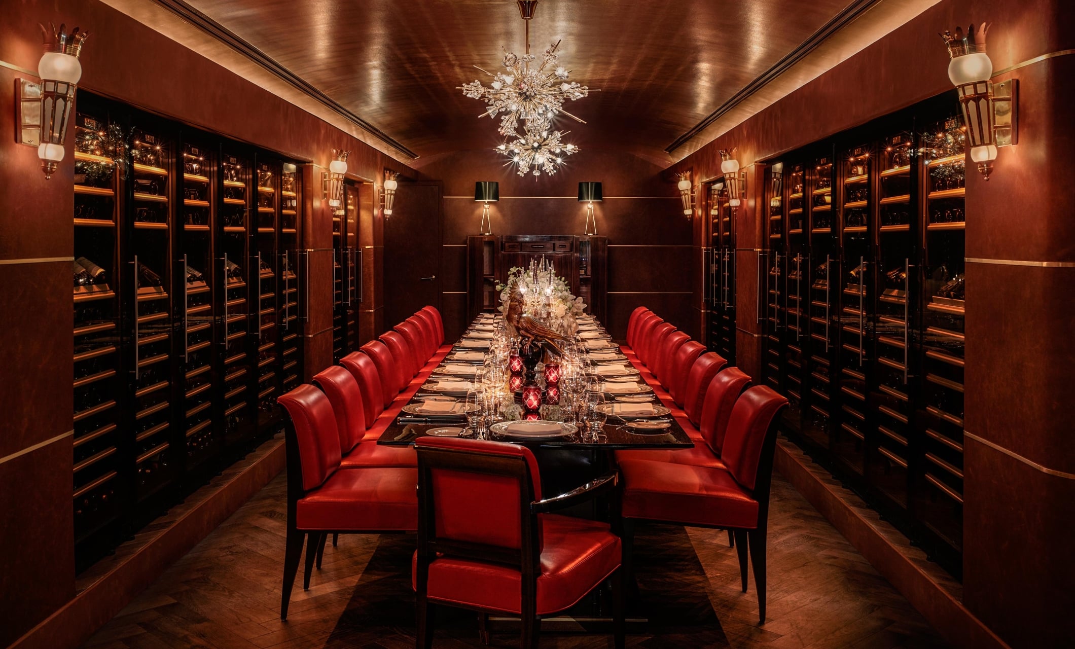 A long dining table set in a wine cellar with red leather chairs and a chandelier.